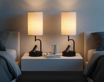 May include: Two matching bedside lamps with cream-colored shades and black bases. Each lamp has a built-in charging station with a phone. A white nightstand holds a mug, glasses, and a book. The lamps are on either side of a bed.