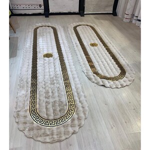 May include: Two oval-shaped, cream-colored rugs with a textured, shaggy surface. Each rug features a prominent black and gold Greek key border pattern and a central gold emblem with the letter E, creating a cohesive set of floor coverings.