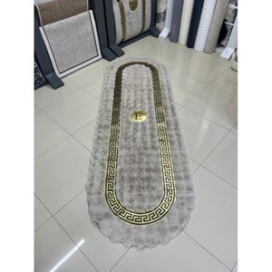 May include: An oval-shaped beige rug with a textured, plush surface. The rug features a prominent gold-colored border with a repeating Greek key pattern and a central gold circular emblem containing the capital letter E in the middle.