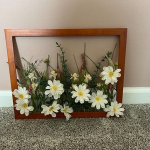 May include: A rectangular wooden frame with a clear center, filled with artificial flowers and greenery. White daisies with yellow centers are prominent, along with other small white and pink flowers and green foliage. The frame is a warm brown color.