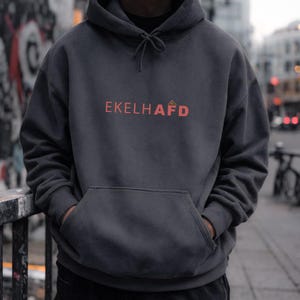 Könnte beinhalten: Dunkelgrauer Kapuzenpullover mit Kordelzug und Vordertasche. Das Wort "EKELHAFD" ist rot auf die Brust gedruckt. Der Hoodie ist aus weichem Material.