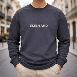 Może przedstawiać: Ciemnoszara bluza z napisem "EKELHAFD" w jasnobeżowym kolorze. Bluza ma długie rękawy i okrągły dekolt. Osoba ma na sobie beżowe spodnie i ręce w kieszeniach. Tło to rozmyta scena uliczna.