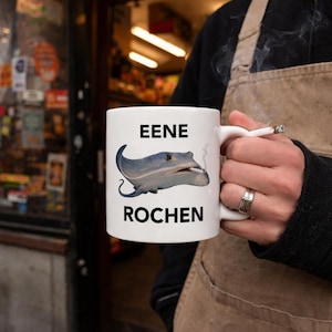 May include: White ceramic mug featuring a cartoon stingray smoking a cigarette, with the words "EENE" and "ROCHEN". The mug is held by a person wearing a brown apron. The mug is a standard size.