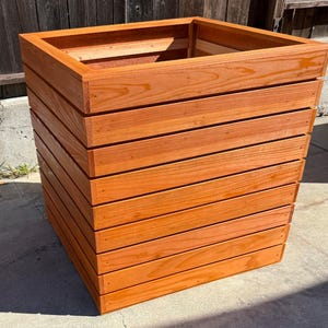Puede incluir: Una jardinera cuadrada de madera con un acabado marrón rojizo natural. La jardinera está construida con listones de madera horizontales, creando un efecto en capas. El borde superior presenta un marco más grueso.
