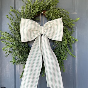 May include: A green wreath with lush foliage and a large bow. The bow is made of fabric with vertical stripes in white and light green. The wreath is hanging on a blue door.