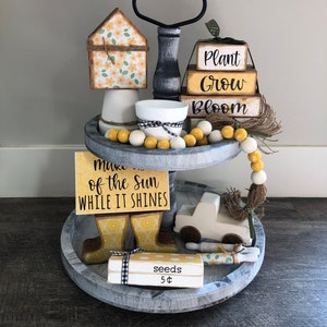 May include: A two-tiered wooden tray with a farmhouse style. The top tier features a small house with a floral pattern, a black and white candle holder, a white candle, and a string of yellow and white beads. The bottom tier features a sign that says "Make the most of the sun while it shines", a pair of yellow rain boots, a white toy truck, a wooden rolling pin with a floral pattern, and a small wooden box that says "seeds 5$".