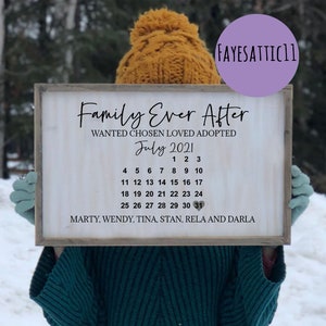 May include: A rectangular wooden sign with a white background and a brown frame. The text reads Family Ever After, WANTED CHOSEN LOVED ADOPTED, July 2021, a calendar grid from 1 to 31 with a heart on the 31st, and the names MARTY, WENDY, TINA, STAN, RELA AND DARLA.
