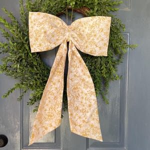 May include: A decorative wreath featuring a lush green base and a large bow. The bow is made of fabric with a cream background and a yellow floral pattern. The wreath is hanging on a blue door.