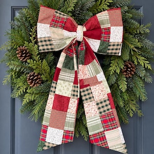 May include: A lush evergreen wreath featuring natural brown pinecones and a large, decorative bow made from a patchwork of fabric squares in red, green, white, and tan colors with various plaid, floral, and solid patterns.