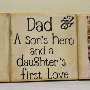 May include: A wooden sign with the text "Dad, A son's hero and a daughter's first love" in black lettering on a beige background. The sign is divided into three sections, with a photo of a man in the left section and a photo of a young boy in the right section.