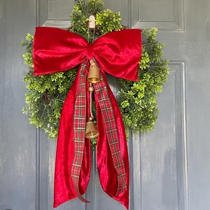 May include: A Christmas wreath featuring a green foliage base, a large red velvet bow, and a red plaid ribbon. Two small gold bells dangle from the bow, enhancing the festive look of this seasonal decoration.
