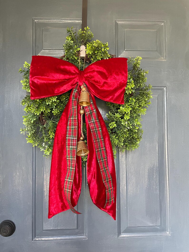 May include: A Christmas wreath featuring a green foliage base, a large red velvet bow, and a red plaid ribbon. Two small gold bells dangle from the bow, enhancing the festive look of this seasonal decoration.
