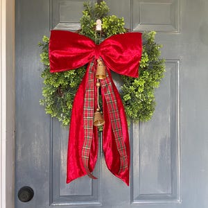 May include: A Christmas wreath featuring a dense green base, a large red velvet bow, and a red and green tartan ribbon. Two small golden bells dangle from the bow. This festive wreath is designed for door decoration, adding a touch of holiday cheer.