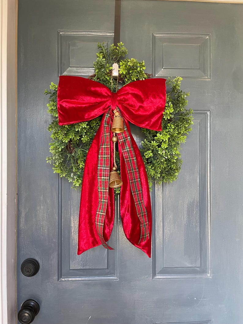 May include: A Christmas wreath featuring a dense green base, a large red velvet bow, and a red and green tartan ribbon. Two small golden bells dangle from the bow. This festive wreath is designed for door decoration, adding a touch of holiday cheer.