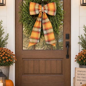 May include: A brown wooden front door featuring a green boxwood wreath adorned with a large plaid bow in shades of orange, yellow, and red. The door has a black handle and lock, and the entryway includes a tan welcome mat and orange pumpkins.