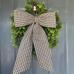 May include: A decorative wreath with a green, leafy base and a large plaid bow. The bow features a pattern of dark green, red, and white checks. The wreath is hanging on a blue door, creating a welcoming aesthetic.