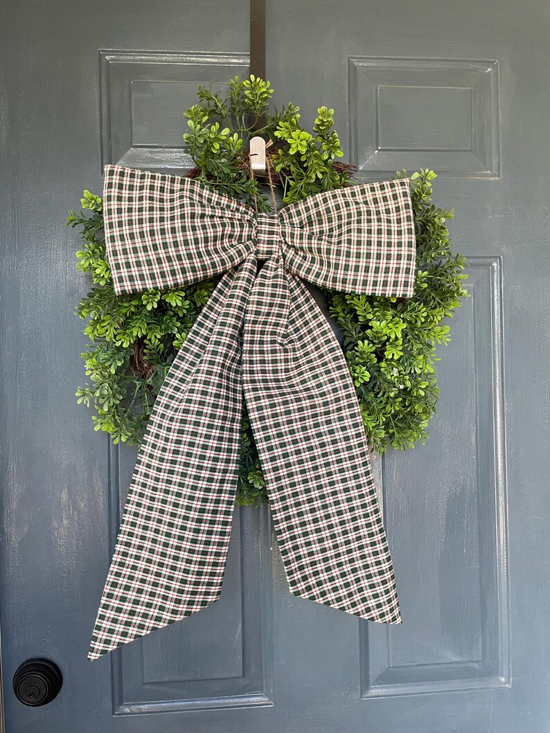 May include: A decorative wreath with a green, leafy base and a large plaid bow. The bow features a pattern of dark green, red, and white checks. The wreath is hanging on a blue door, creating a welcoming aesthetic.