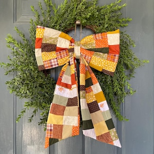 May include: A large decorative bow made of patchwork fabric featuring various autumn-inspired patterns in shades of orange, yellow, brown, green, and cream. The bow has two long, trailing tails and is attached to a green leafy wreath.
