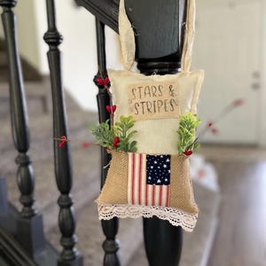 Puede incluir: Adorno colgante decorativo con el texto "STARS & STRIPES". Presenta una bandera estadounidense sobre arpillera, con vegetación y bayas rojas. El borde inferior está adornado con encaje blanco. Cuelga de un poste negro.