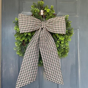 May include: A green wreath with a large plaid bow featuring a pattern of green, red, and white checks. The wreath is hanging on a gray door, creating a welcoming and festive display.