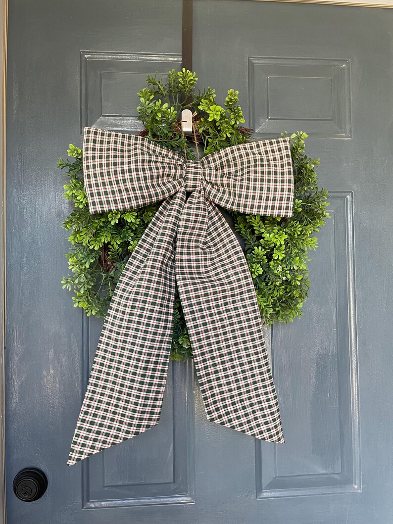 May include: A green wreath with a large plaid bow featuring a pattern of green, red, and white checks. The wreath is hanging on a gray door, creating a welcoming and festive display.