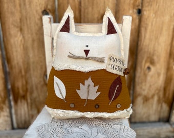 May include: A handmade cat-shaped pillow with a cream-colored face, brown ears, and a mustard-colored body adorned with leaf appliqués. The pillow has the text "PUMPKIN SEASON" and is sitting on a white chair with a crocheted doily.