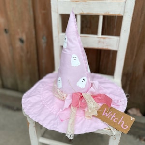 May include: A pink witch hat with white ghost designs and a wide brim sits on a white chair. The hat has a matching pink ribbon and a tag that says "Witch". The hat is a festive Halloween decoration.
