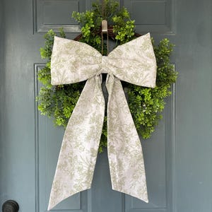 May include: A decorative wreath with a lush green base and a large bow. The bow features a white fabric with a light green floral pattern. The wreath is hanging on a blue-green door.
