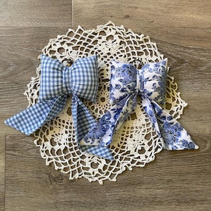 May include: Two fabric bow ornaments with metal rings at the top. One bow features a blue and white gingham check pattern, while the other displays a blue and white floral toile print. Both bows have long, pointed tails extending downwards.