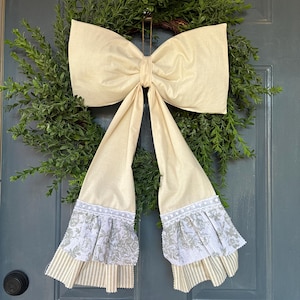 May include: A decorative wreath with a green leafy base and a large, cream-colored bow. The bow has long, flowing tails with floral patterned and striped fabric accents. The wreath is hanging on a blue door.