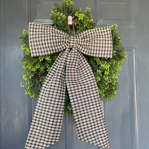 May include: A decorative wreath featuring a lush green base and a large plaid bow. The bow has a pattern of dark green, red, and white squares. The wreath is hanging on a blue door.