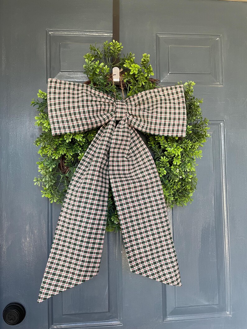 May include: A decorative wreath featuring a lush green base and a large plaid bow. The bow has a pattern of dark green, red, and white squares. The wreath is hanging on a blue door.