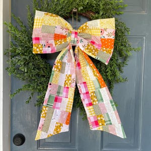 May include: A colourful patchwork bow adorns a green foliage wreath. The bow is made from patterned fabrics in pink, yellow, orange, and green. This decorative wreath provides a bright and cheerful accent for a door or wall, adding a touch of handmade charm.