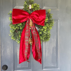 May include: A Christmas wreath featuring a green base, a large red velvet bow, and a red plaid ribbon. Two small gold bells dangle from the bow, creating a traditional festive decoration for the holiday season.