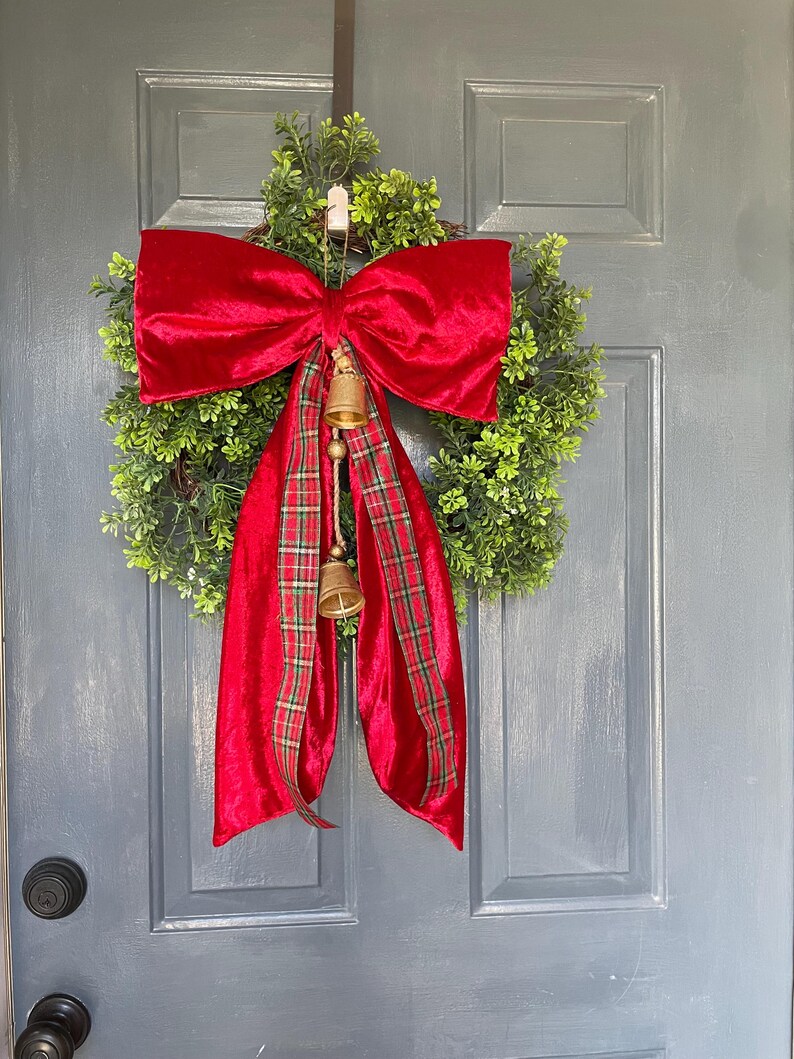 May include: A Christmas wreath featuring a green base, a large red velvet bow, and a red plaid ribbon. Two small gold bells dangle from the bow, creating a traditional festive decoration for the holiday season.