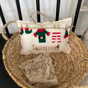 Könnte beinhalten: Ein dekoratives Weihnachtskissen mit Filz-Elfenkleidung und dem Schriftzug "NORTH POLE". Das Kissen ist mit Miniatur-Elfen-Schuhen, einer Jacke, einer Hose und einem Hut aus Filz verziert. Das Kissen liegt in einem geflochtenen Korb.