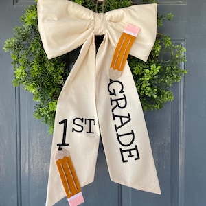 May include: A white fabric bow with a large bow on top and two long tails. The tails have the words "1st" and "GRADE" printed on them. There are two felt pencils attached to the tails. The bow is hanging on a green wreath.