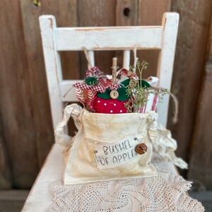 May include: A rustic, cream-colored fabric bag labeled "BUSHEL OF APPLES" filled with decorative apple ornaments and greenery. The bag is adorned with a small bell and sits on a white, distressed chair with a lace doily.