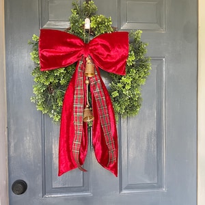 May include: A festive Christmas wreath featuring a full green base, a large red velvet bow, and a plaid ribbon. Two small gold bells hang below the bow. This door decoration is perfect for the holiday season.
