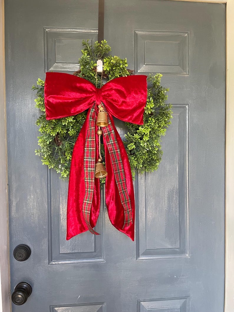 May include: A festive Christmas wreath featuring a full green base, a large red velvet bow, and a plaid ribbon. Two small gold bells hang below the bow. This door decoration is perfect for the holiday season.