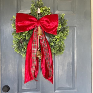 May include: A Christmas wreath featuring a large red velvet bow and red plaid ribbon. The wreath is made of green foliage and decorated with two small gold bells. It hangs on a grey door.