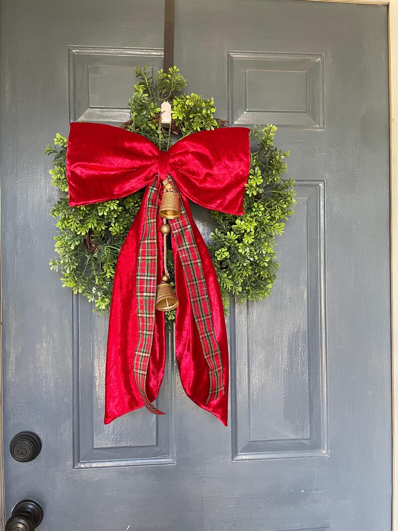 May include: A Christmas wreath featuring a large red velvet bow and red plaid ribbon. The wreath is made of green foliage and decorated with two small gold bells. It hangs on a grey door.