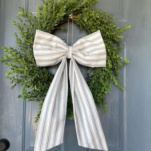 May include: A green, leafy wreath with a large bow made of cream-colored fabric with blue stripes. The bow is centered on the wreath and hangs on a blue door. The wreath is a decorative item for the home.