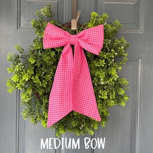 May include: A round green wreath with a vibrant pink plaid bow. The bow is centered on the wreath and has long, flowing tails. The text "MEDIUM BOW" is displayed below the wreath.