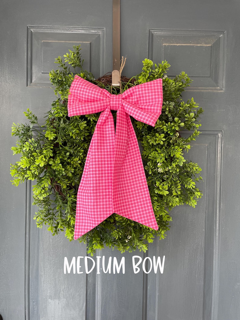 May include: A round green wreath with a vibrant pink plaid bow. The bow is centered on the wreath and has long, flowing tails. The text "MEDIUM BOW" is displayed below the wreath.