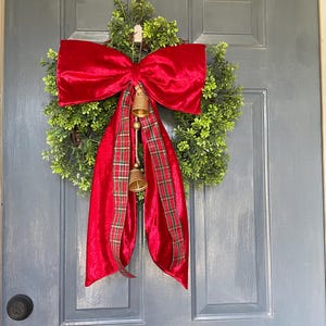 May include: A Christmas wreath featuring a green base, a large red velvet bow, and a red plaid ribbon. Two golden bells dangle from the bow, enhancing the festive look. The wreath is hung on a grey door, ready for the holidays.