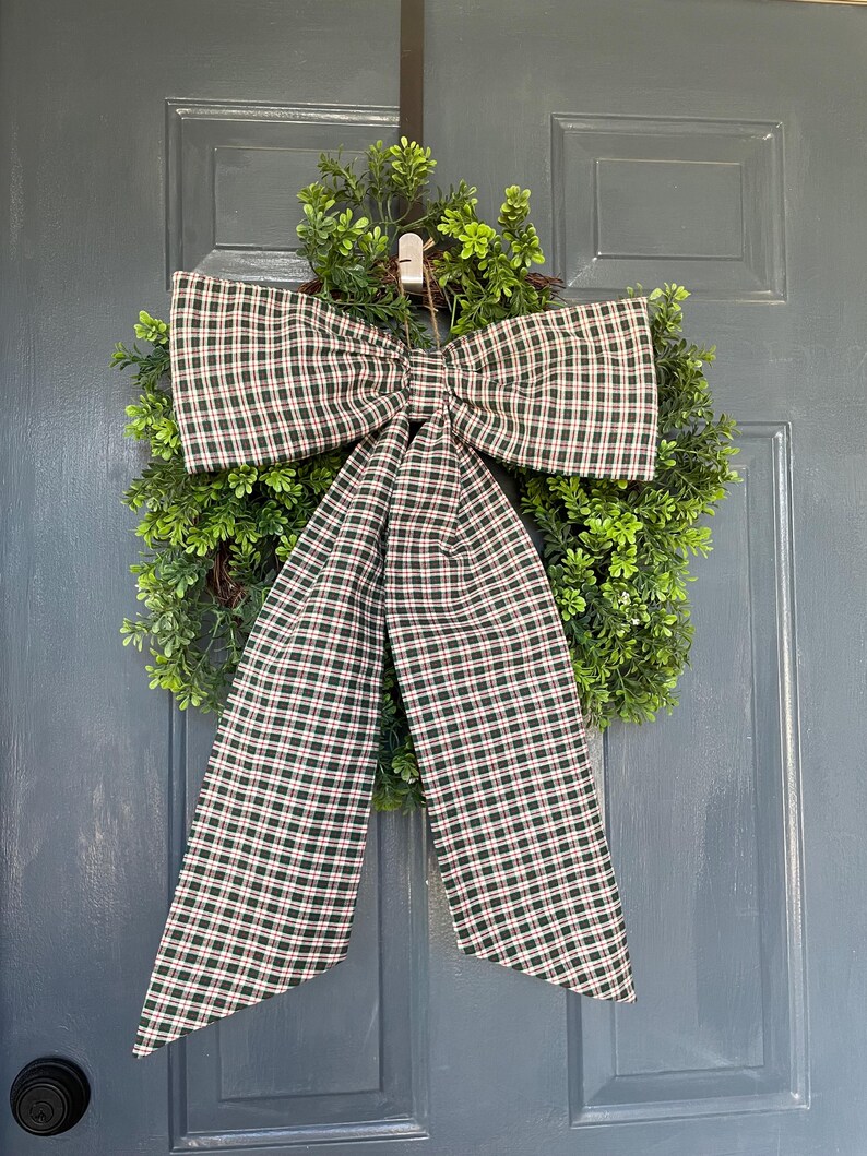 May include: A green wreath with a large plaid bow, featuring a pattern of dark green, red, and white checks. The wreath is made of green foliage and hangs on a gray door, creating a welcoming aesthetic.