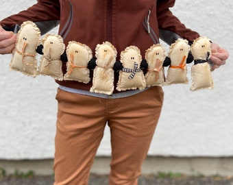 May include: A garland of handmade ghost dolls. Each doll is made of beige fabric with a ruffled edge, black button eyes, and a stitched mouth. The dolls have black or orange accents, including scarves and twine. The garland is held up against a white wall.