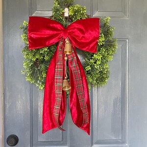 May include: A Christmas wreath featuring a green base, a large red velvet bow, and a plaid ribbon. Two small gold bells hang from the bow. This festive decoration is perfect for the holiday season.