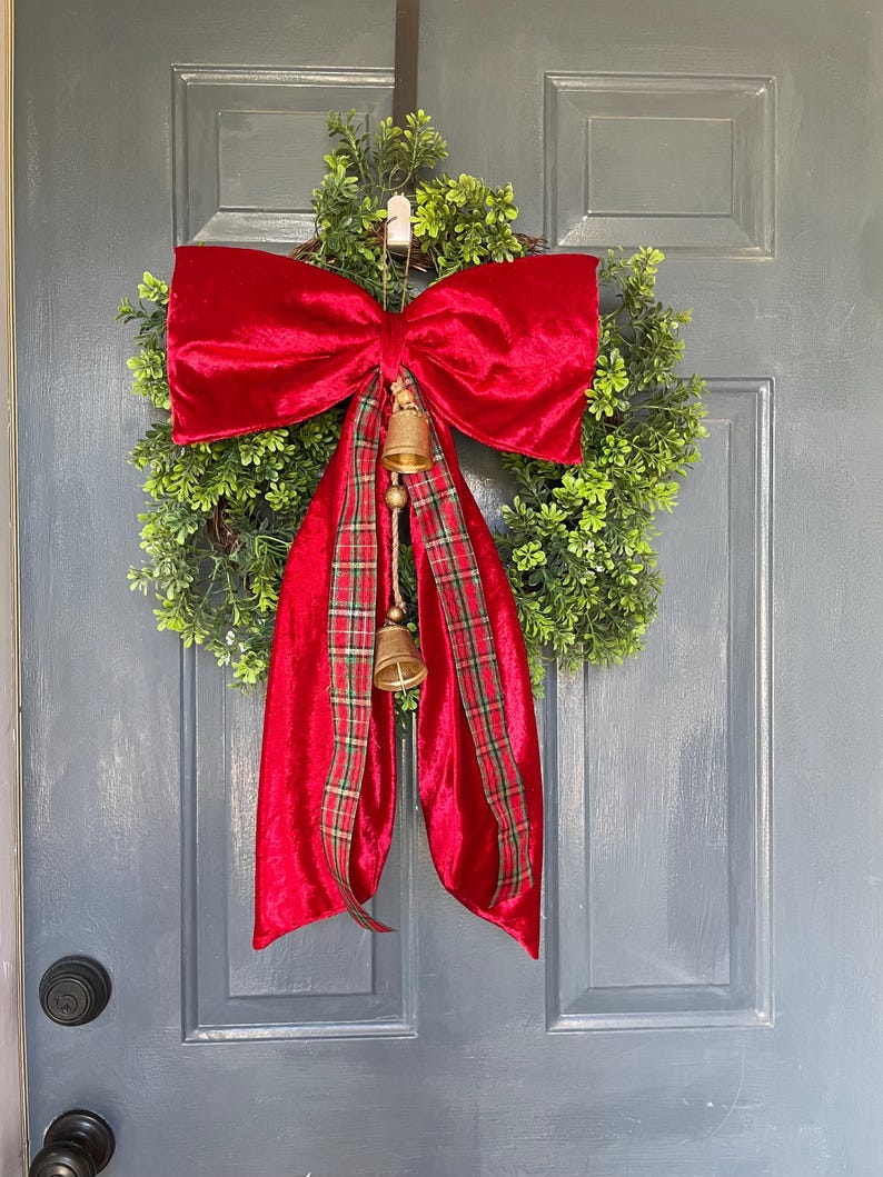 May include: A Christmas wreath featuring a green base, a large red velvet bow, and a plaid ribbon. Two small gold bells hang from the bow. This festive decoration is perfect for the holiday season.